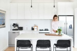 In a modern white Cremorne apartment kitchen adorned with pendant lights, an island, and black chairs, Emma Blomfield arranges items on a wooden tray next to a potted plant and bottles of sparkling water.