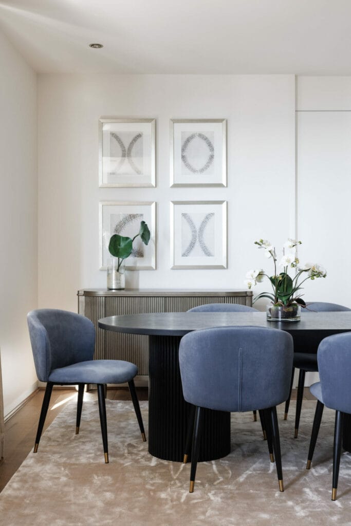 A modern, luxe dining room features a black round table surrounded by four blue upholstered chairs, framed abstract art on the wall, and indoor plants adorning both the credenza and the table.