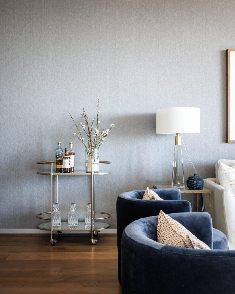 In a modern living room exuding CITY LUXE, a small bar cart adorned with bottles, glassware, and a vase of branches stands beside two dark blue velvet armchairs and a floor lamp.