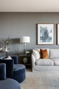 In a city luxe modern living room, a light grey sofa adorned with brown and white pillows pairs seamlessly with blue velvet chairs, a side table graced by a lamp, and abstract artwork on the grey wall; Emma Blomfield sits comfortably within this stylish space.