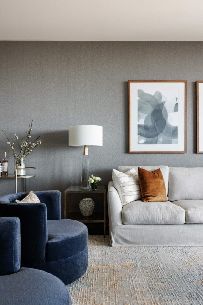In a city luxe modern living room, a light grey sofa adorned with brown and white pillows pairs seamlessly with blue velvet chairs, a side table graced by a lamp, and abstract artwork on the grey wall; Emma Blomfield sits comfortably within this stylish space.