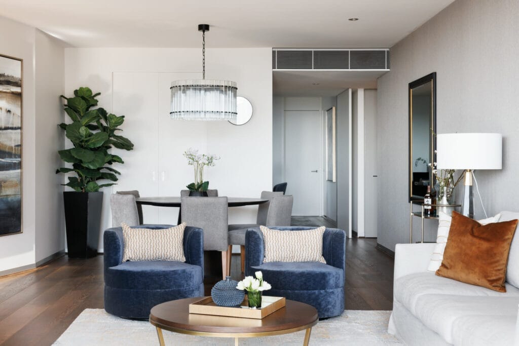 A modern living room in this luxurious city unit features a sofa with brown cushions, two blue armchairs, a round coffee table, a dining table with grey chairs, and a potted plant, all beautifully complemented by wall art and a ceiling light fixture to complete the stylish space.