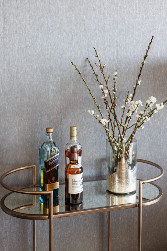 A small metal and glass table from the LUXE collection, holding three bottles of whiskey and a vase with white flowering branches, adds an elegant touch to any city dwelling, with Emma Blomfield standing in front of a plain grey wall.