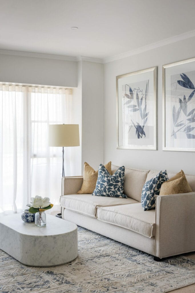 In the York St Unit's contemporary living room, a blonde woman named Emma Blomfield sits on a beige sofa adorned with blue and beige cushions, facing a white marble coffee table on a patterned rug, with a floor lamp nearby and two framed botanical prints on the wall.