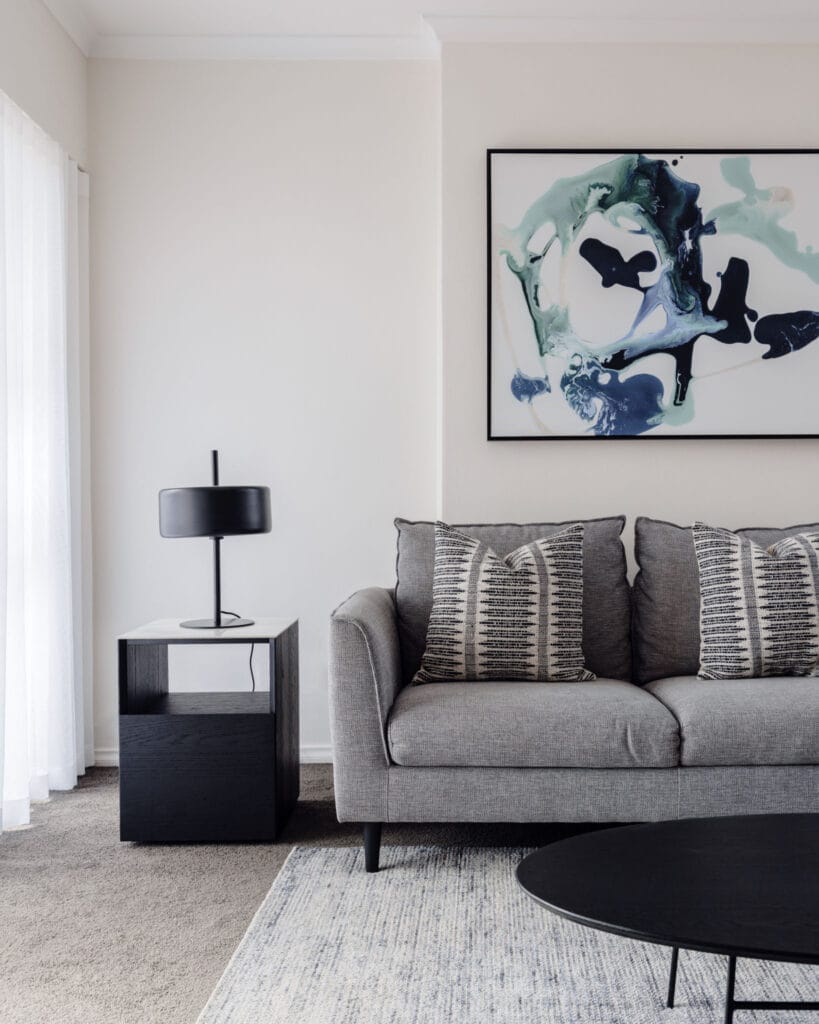 A modern living room features a grey sofa adorned with two patterned cushions, a black side table paired with a matching lamp, abstract wall art, and a round black coffee table in front of a large window dressed in white curtains.
