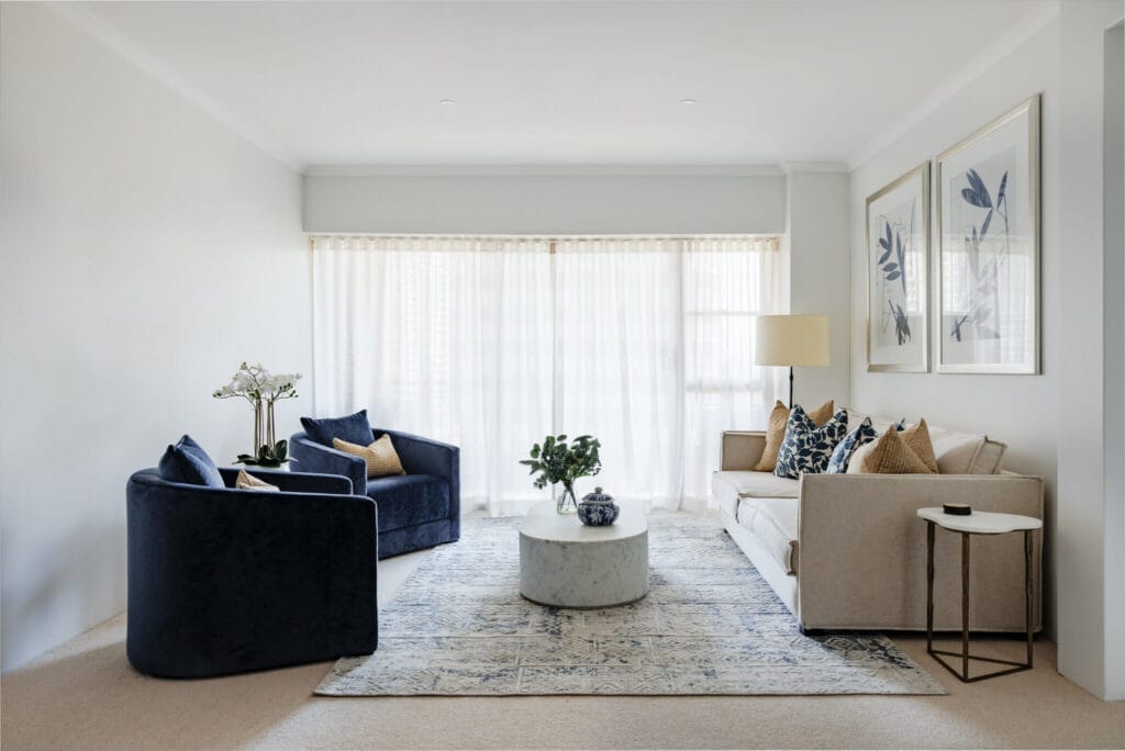 In a modern living room featuring a beige sofa, navy armchairs, a round marble coffee table, and large windows with sheer curtains, the space is elegantly decorated with framed artwork, plants, and neutral-toned furnishings.