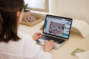 Emma Blomfield is seated at her desk, immersed in crafting budget-friendly designs on a laptop with interior design software, accompanied by a pink potted plant and a wicker basket.