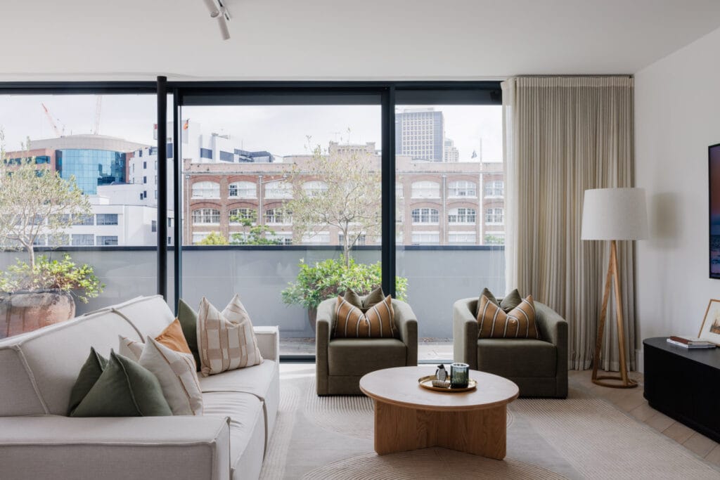 A modern living room featuring a white sofa, two green armchairs, and a round wooden coffee table creates the perfect setting for hosting Easter or infusing the space with Easter-themed décor to complement the cityscape views through expansive windows.
