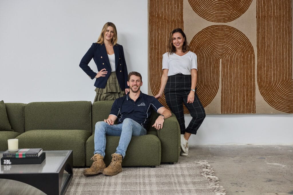Emma Blomfield and another woman stand smiling behind a man seated on a green sofa in a modern living room designed by EB Studios, featuring eco-friendly elements, with a large abstract artwork on the wall and a coffee table in the foreground.