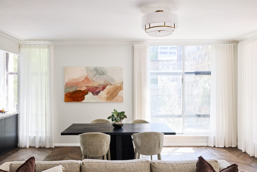 A modern dining area featuring a black table, four beige chairs, a potted plant centerpiece, abstract art on the wall, and large windows with sheer white curtains is perfect interior design inspiration for a cozy winter feel—and when Emma Blomfield is in the image, her presence adds an inviting touch.