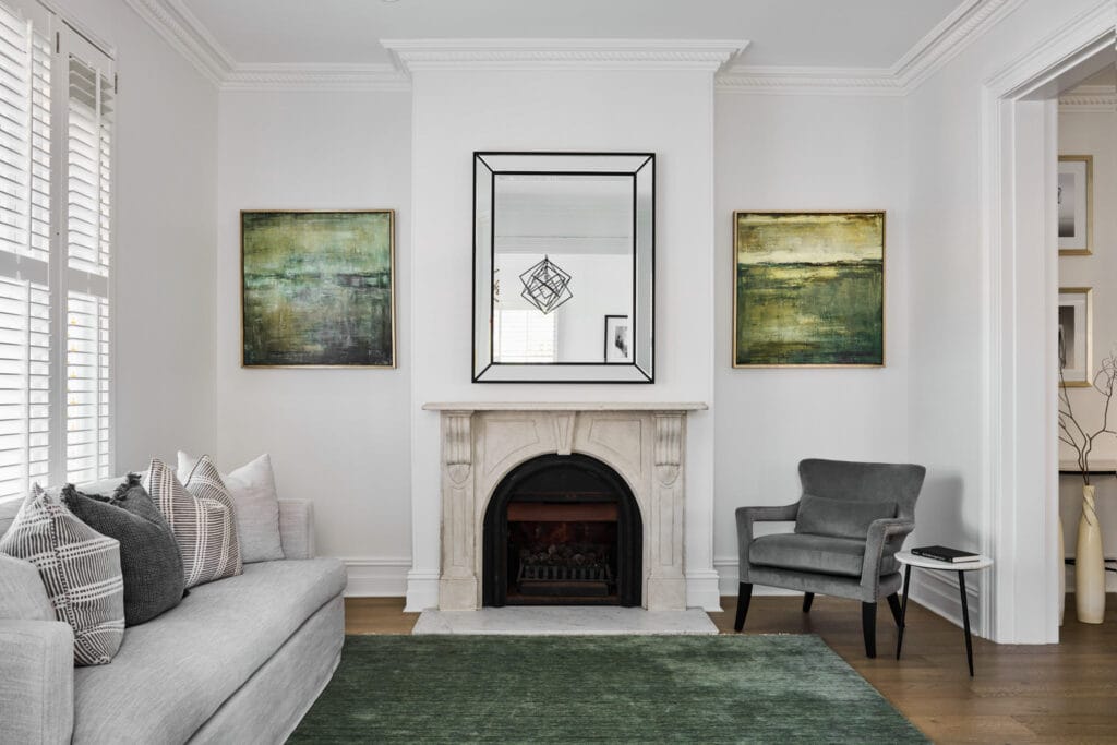 A modern living room featuring a gray sofa and armchair, a green rug, and a marble fireplace is beautifully highlighted by a large mirror and two abstract green paintings on the white wall.