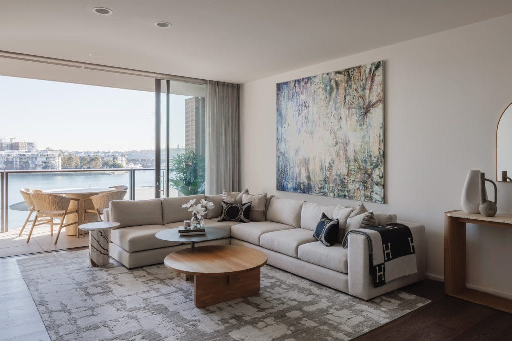 A spacious living room features a large sectional, wooden coffee table, abstract art, and floor-to-ceiling windows with stunning water views over the Barangaroo Terrace balcony.