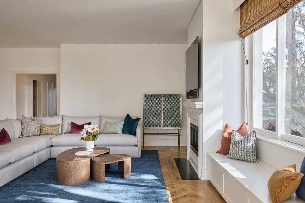 This modern living room features a light gray sectional, colorful cushions, nested wooden tables, a blue rug, TV, and large sunny windows.