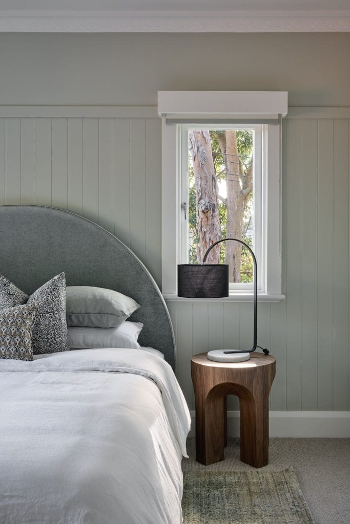 A neatly made bed with a gray upholstered headboard and patterned pillows sits by a wooden nightstand and window overlooking a tree.