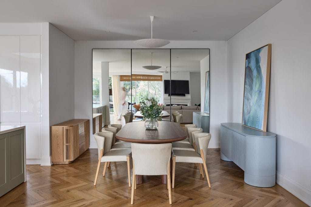A stylish dining room features a wooden oval table, eight cream chairs, a large wall mirror, and chic decor like flowers and art.
