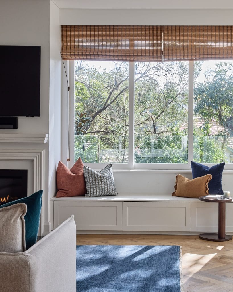 A sunlit living room features a window seat, colorful cushions, round table, and fireplace beside a large window with garden views.