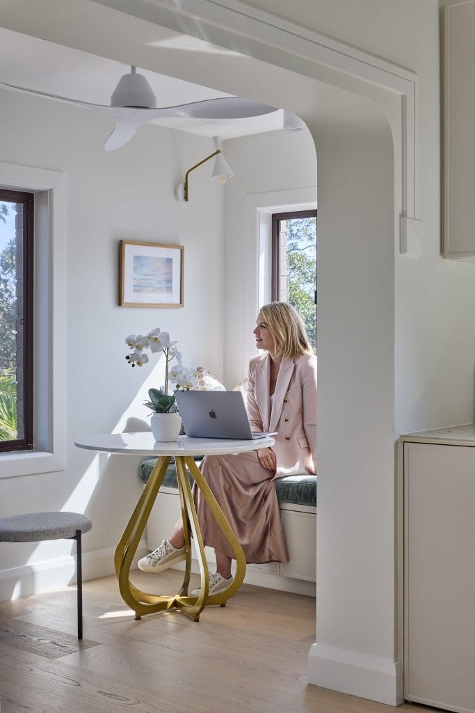 Emma Blomfield, dressed in a pink suit, works on her laptop at a cozy table in a sunny nook with plants and wall art.