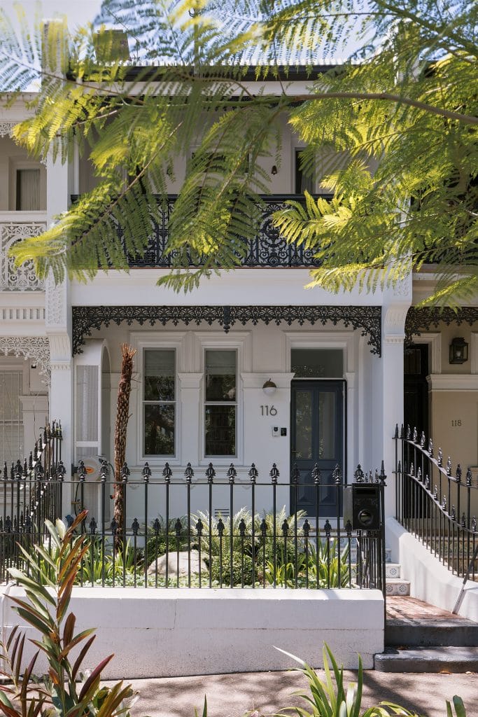 A white two-story Paddington terrace with a black iron fence, ornate balcony, and dark door sits amid lush greenery and dappled shade.