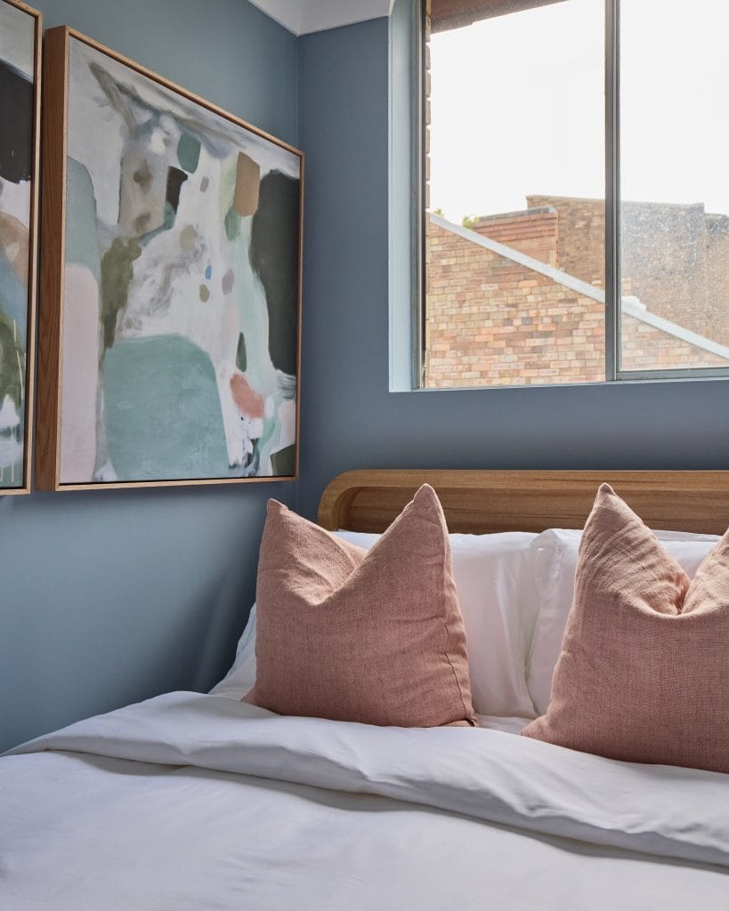 A tidy bed with pink pillows and white sheets sits in a Surry Hills unit with blue walls, art, and a window facing a brick building.