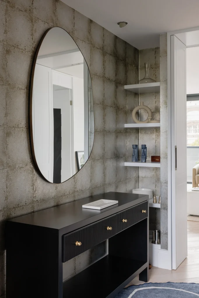 A sleek entryway features a large oval mirror above a black console, white corner shelves with decor, and textured wallpaper.