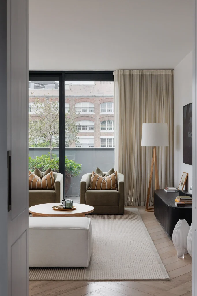 A bright, neutral-toned living room features two armchairs, a round coffee table, TV, floor lamp, and a balcony with potted plants.
