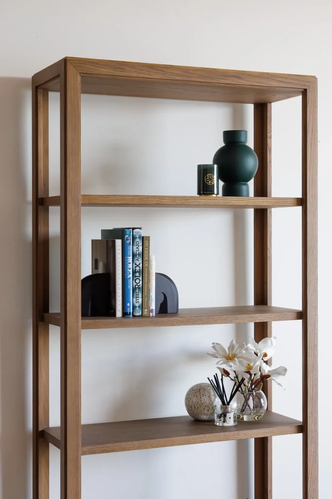The wooden bookshelf displays a green vase and candle on top, books in the middle, and flowers with a decorative sphere below.