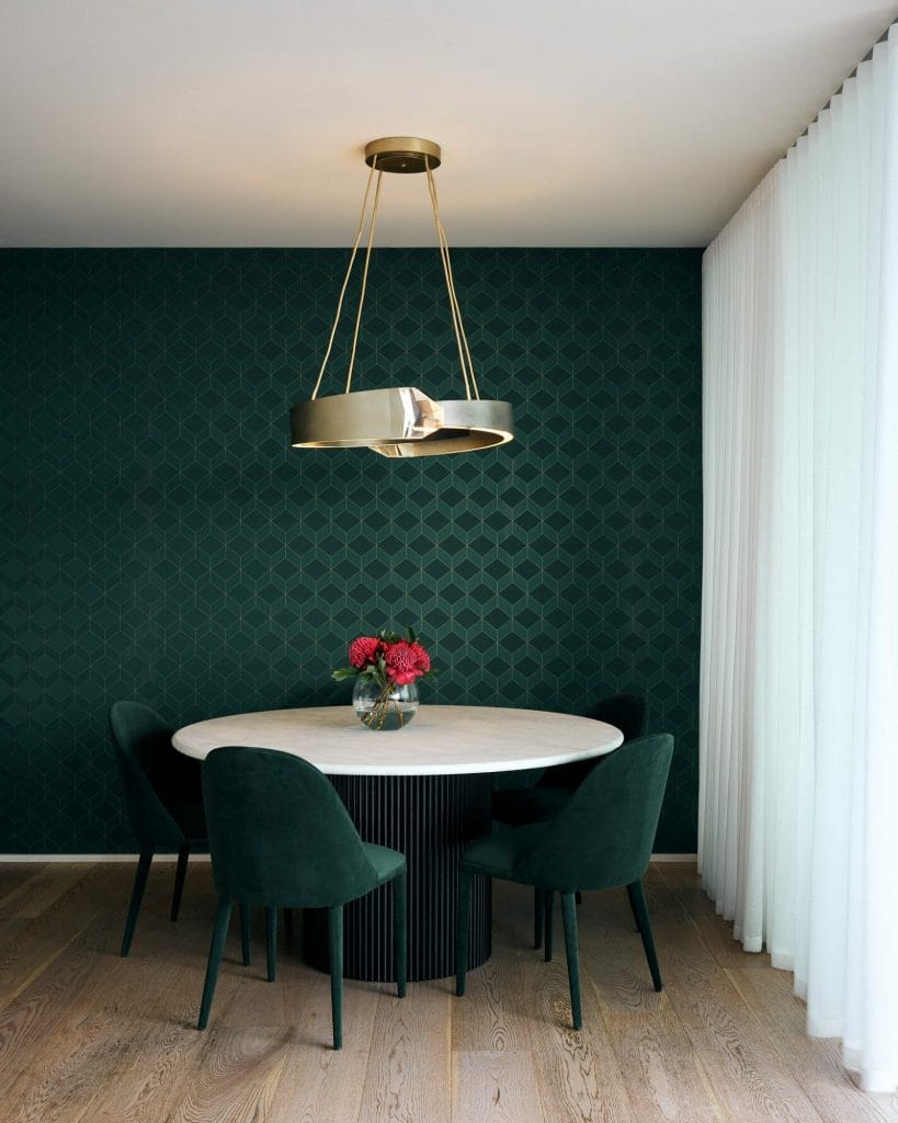 A round dining table with four dark green chairs, red flowers in a vase, and a gold chandelier sit before a dark patterned wall and white curtains.
