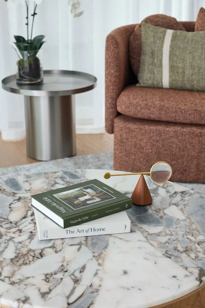 A marble coffee table holds two books and a decorative magnifying glass, with a brown sofa, green cushion, and a potted plant nearby.