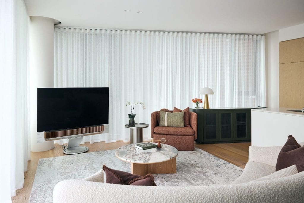 A modern living room featuring a big TV, round marble coffee table, textured rug, plush chairs, and sheer curtains on tall windows.
