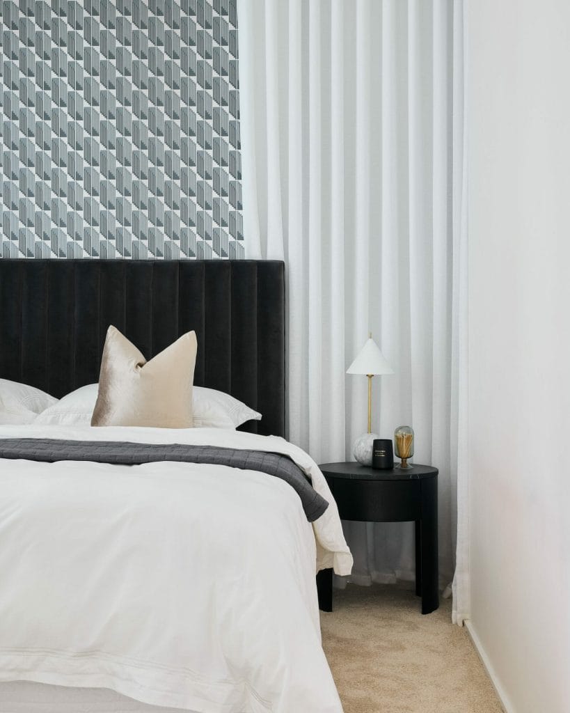 A neatly made bed with white bedding sits in a modern bedroom, featuring a black headboard, geometric accent wall, and nightstand with a lamp and glass.
