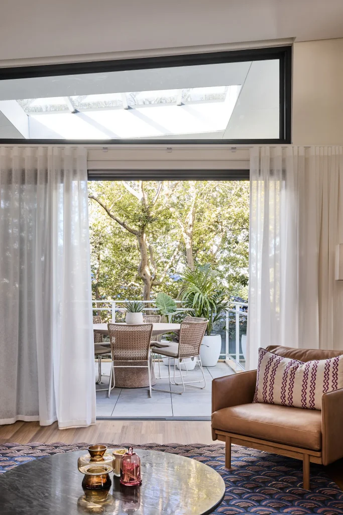 Sheer curtains in the living room reveal a balcony with a table, chairs, potted plants, and views of greenery through a skylight.