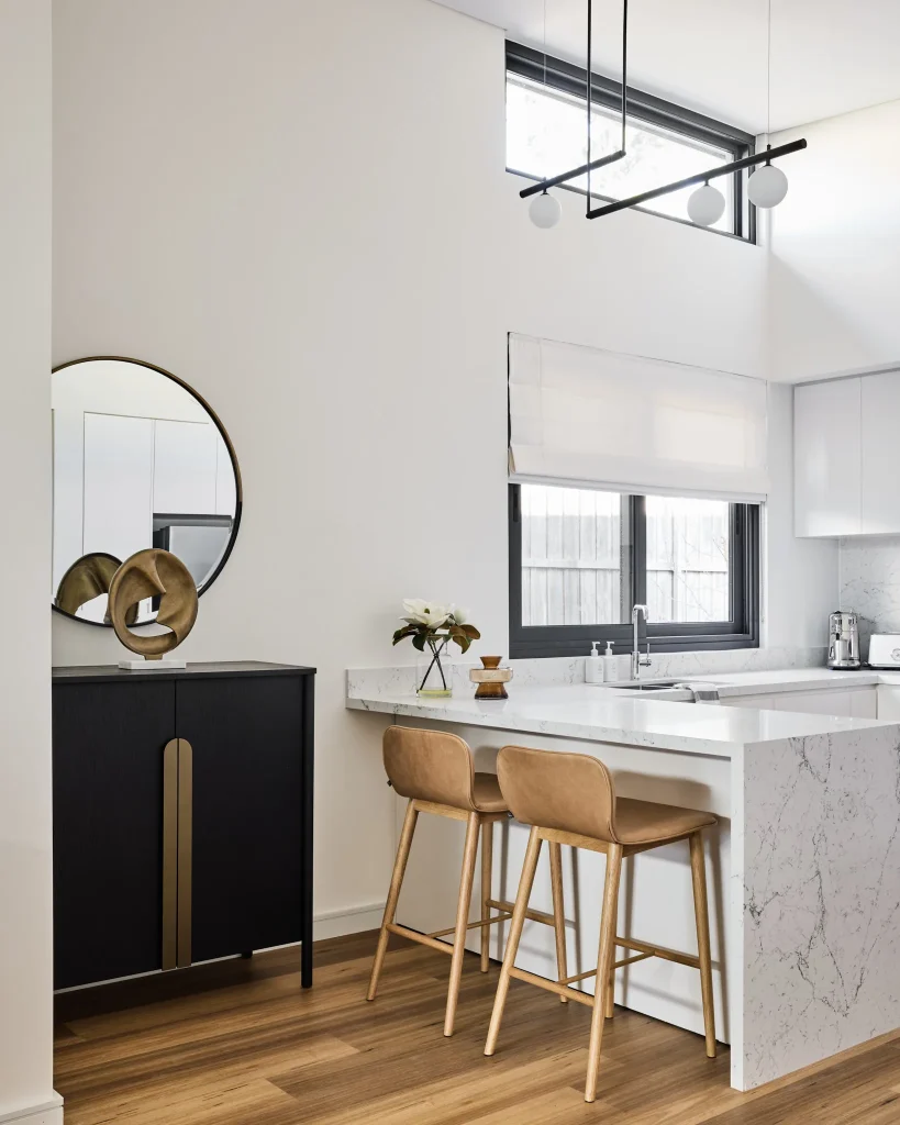 A kitchen features white cabinets, a marble countertop, two tan barstools, a black side cabinet with round mirror, and a window.