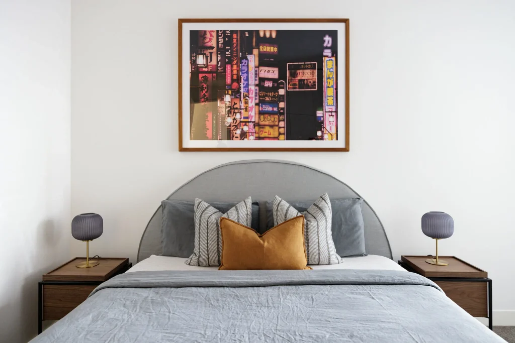 A stylish bedroom features a gray, neatly made bed with pillows, nightstands with lamps, and a city lights photo above the headboard.