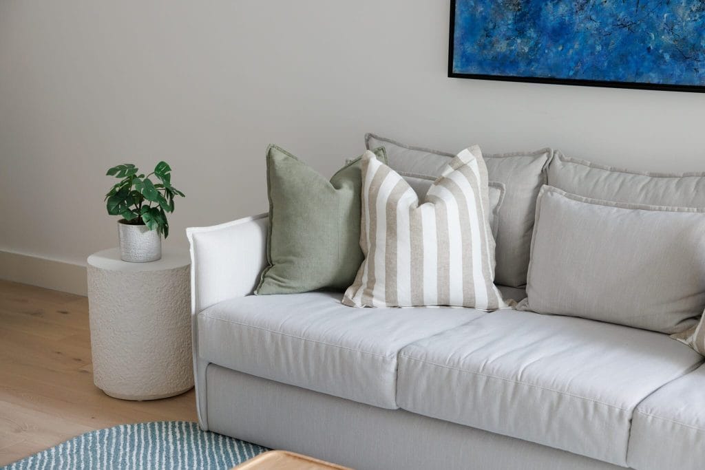 A light gray sofa with assorted pillows sits by a white side table with a potted plant, topped by a blue abstract painting.