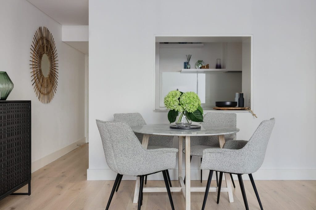 EB Studio’s modern dining area has a round table, four gray chairs, green hydrangeas, and a sleek pass-through in a minimalist room.