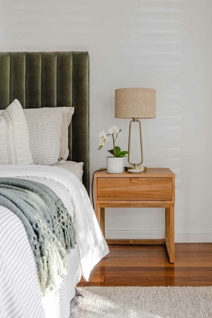 A bed with striped bedding and a green headboard sits next to a wooden nightstand holding a lamp, plant, and books on hardwood floors.