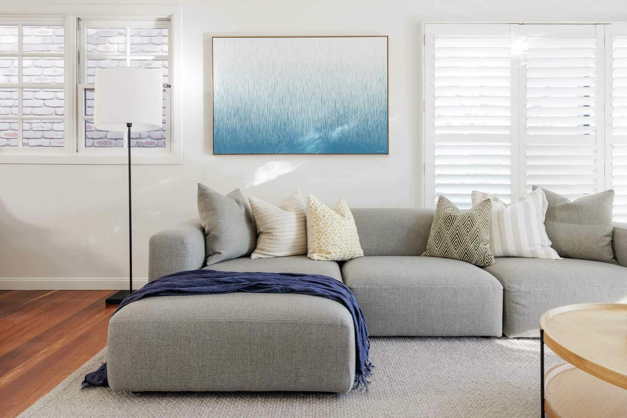 A stylish living room features a light gray sectional with mixed pillows, a blue throw, floor lamp, abstract art, and shuttered windows.