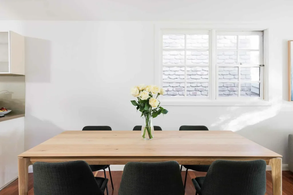 In a sunlit room with white walls and a window, six dark chairs surround a light wood table topped with white roses in glass.