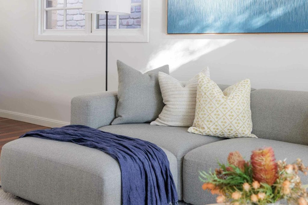 A light gray sectional with two patterned pillows and a dark blue throw sits by a floor lamp under a window and abstract painting, with flowers in front.