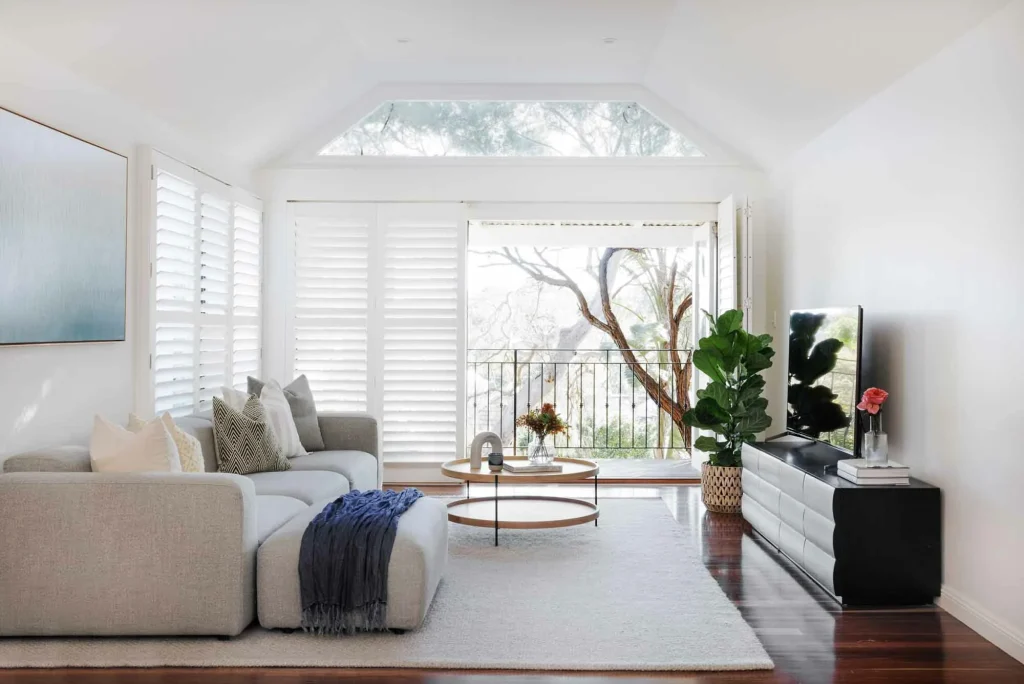 A stylish living room by EB Studio features a gray sectional, round coffee table, TV console, and large windows opening to a balcony with trees.