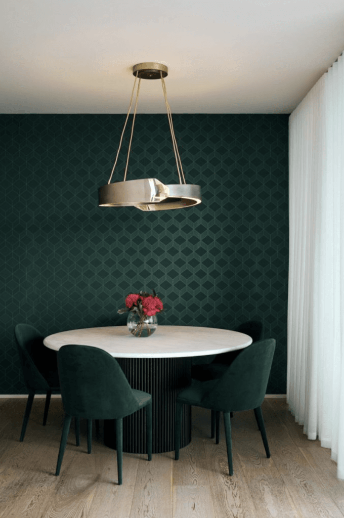 A white round dining table with four dark green chairs on light wood floors sits under a gold chandelier, beside a green accent wall.