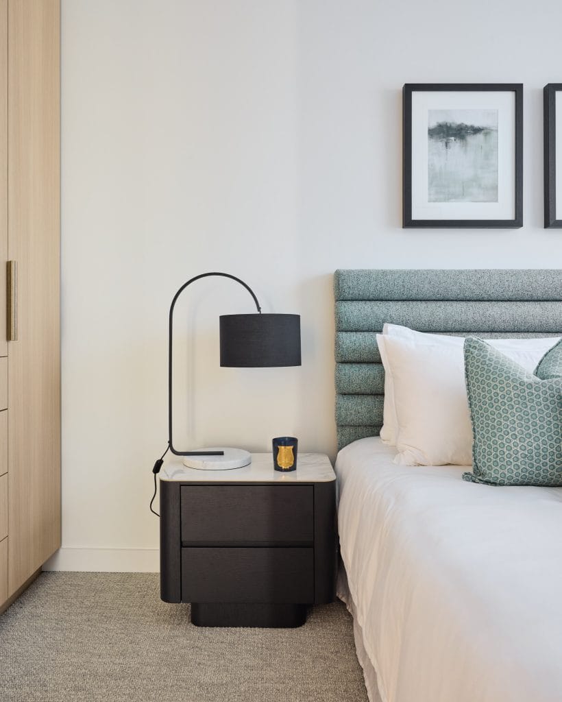 A modern bedroom features a bed with a patterned pillow, black lamp, candle, and two framed artworks on a crisp white wall.