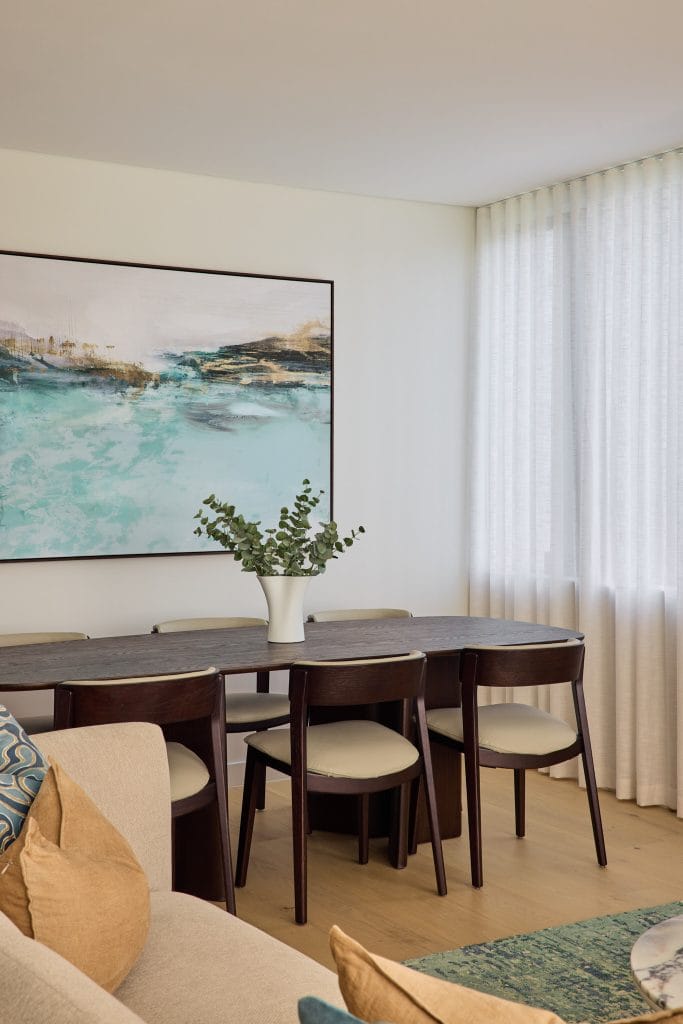 A dining space features a dark wood table, six chairs, a vase of greenery, abstract art, and sheer curtains letting in natural light.