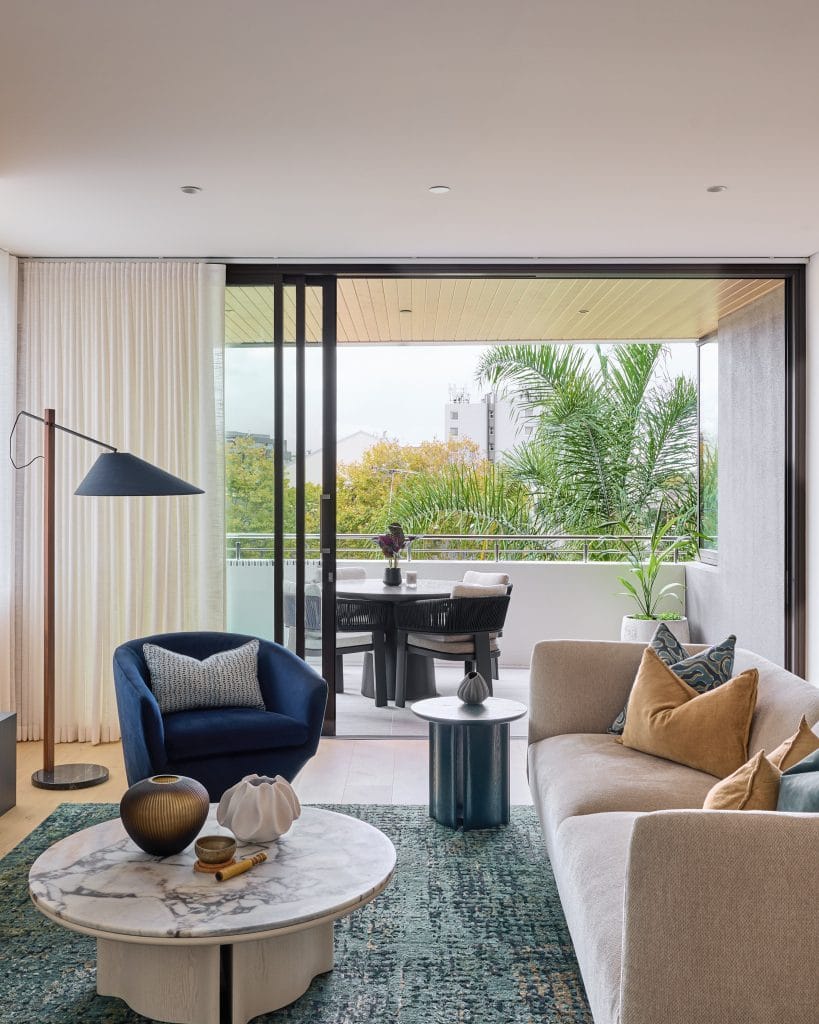 A beige sofa, blue armchair, and marble coffee table decorate a modern living room that opens to a lush balcony with outdoor seating.