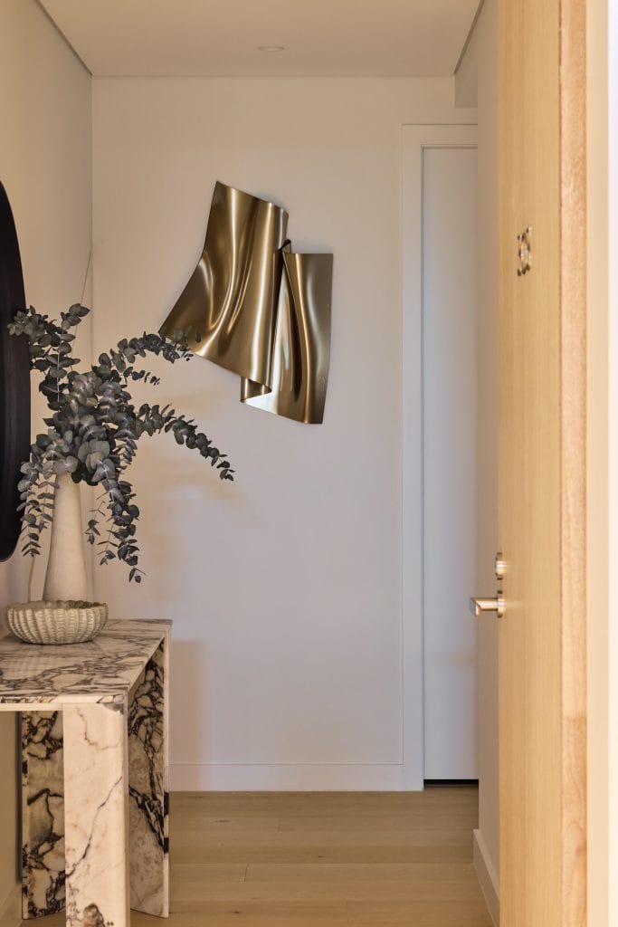 A marble console table with a textured vase of eucalyptus sits beneath a gold abstract wall sculpture in this modern hallway.
