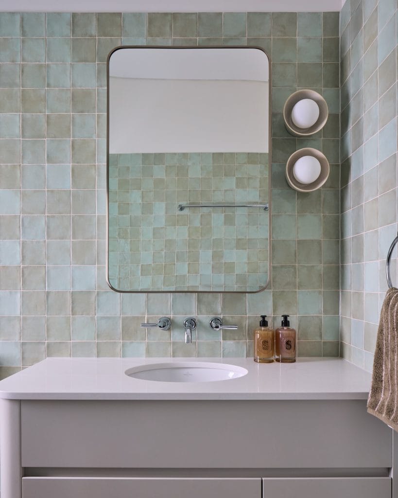 A stylish Mosman bathroom features green square tiles, a rectangular mirror, wall-mounted faucet, shelves, and a brown towel.
