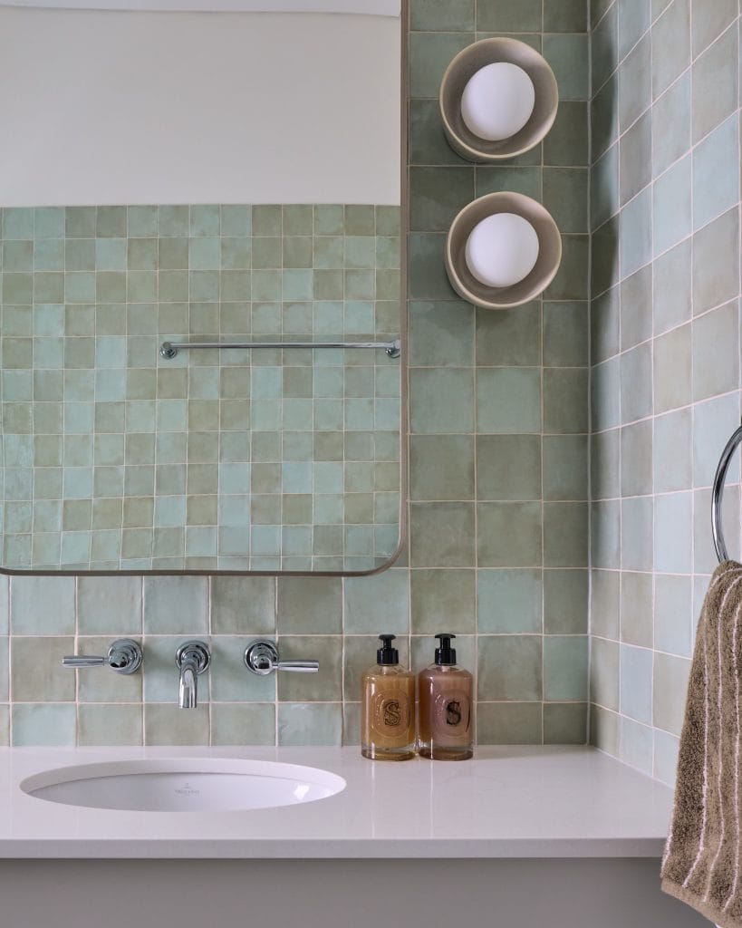 A bathroom with green square tiled walls features a large mirror, wall-mounted faucets, two soap dispensers, and a towel on a ring.