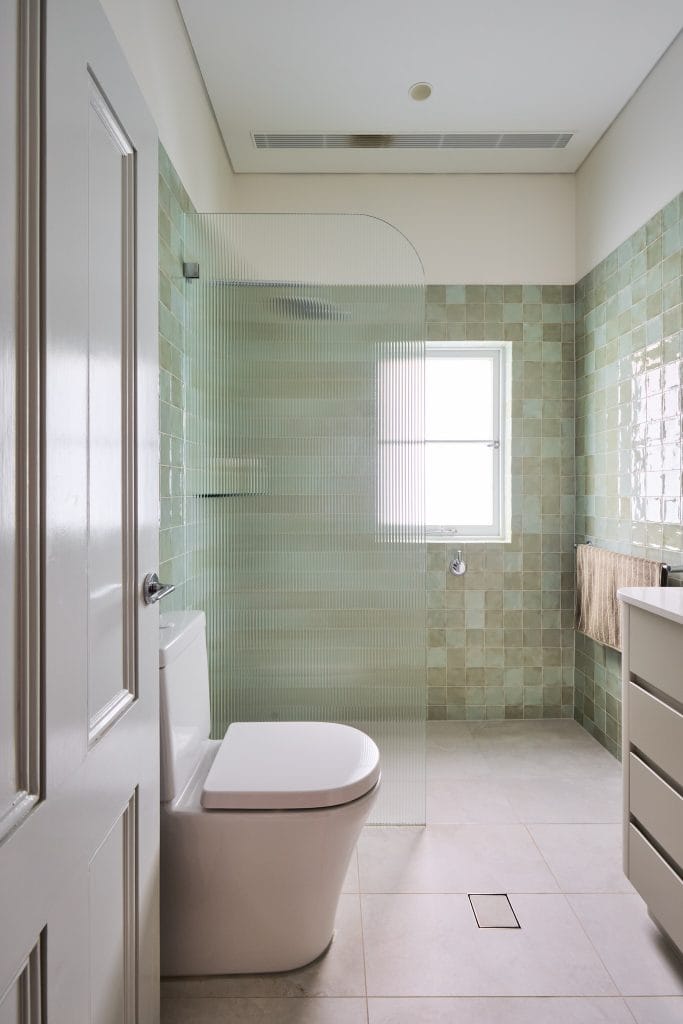 Sleek Mosman bathroom with light green tile walls, white toilet, glass shower divider, window, and modern white vanity.