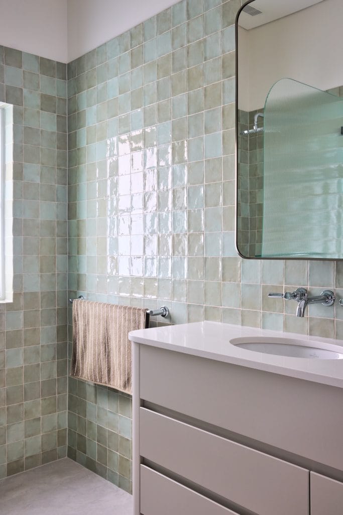 A modern bathroom features light green square tiles, a white vanity with drawers, a striped towel, and a wall mirror above the sink.