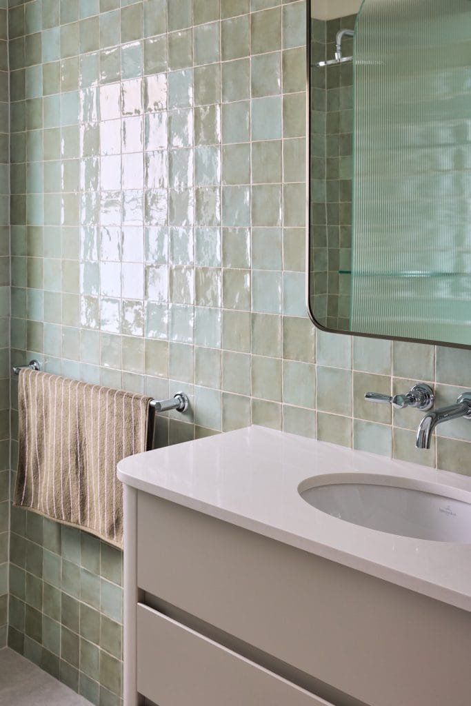 A stylish bathroom features green square tiles, a white vanity, wall-mounted faucet, rectangular mirror, and a striped towel on a rack.